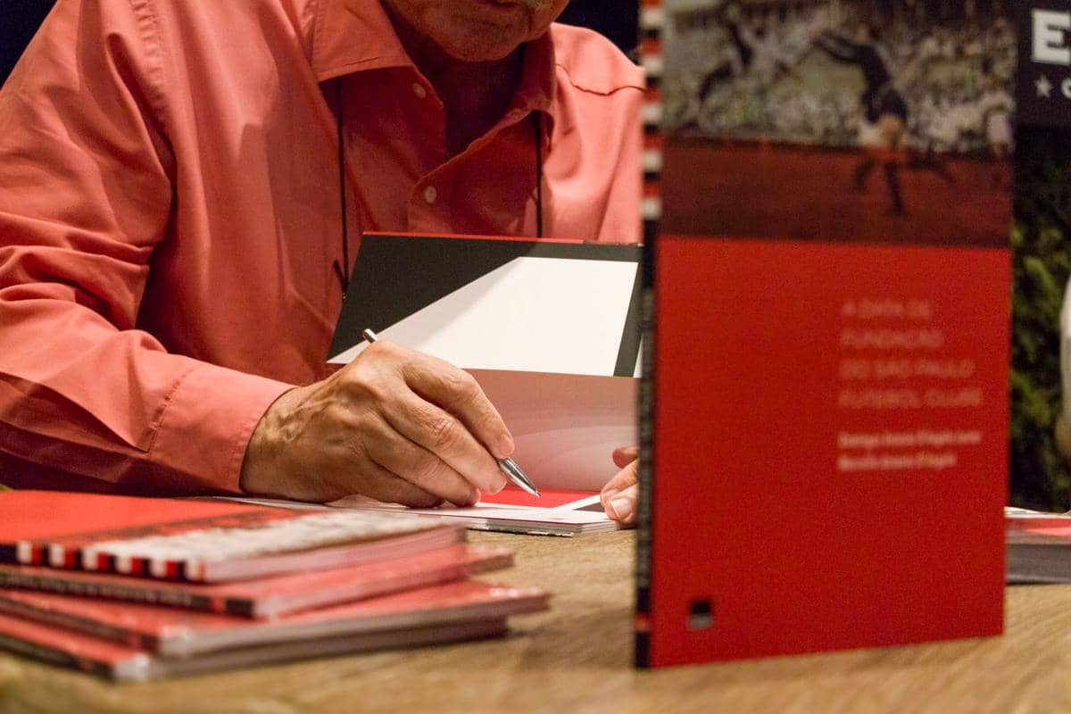 Seorang pria mengenakan kemeja merah muda sedang menandatangani buku di atas meja dengan beberapa buku berwarna merah di sekitarnya.