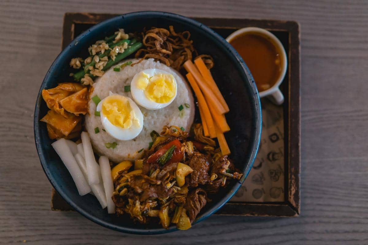 Semangkuk bibimbap berisi nasi, telur rebus setengah matang, sayuran segar, dan daging tumis disajikan di atas meja kayu.
