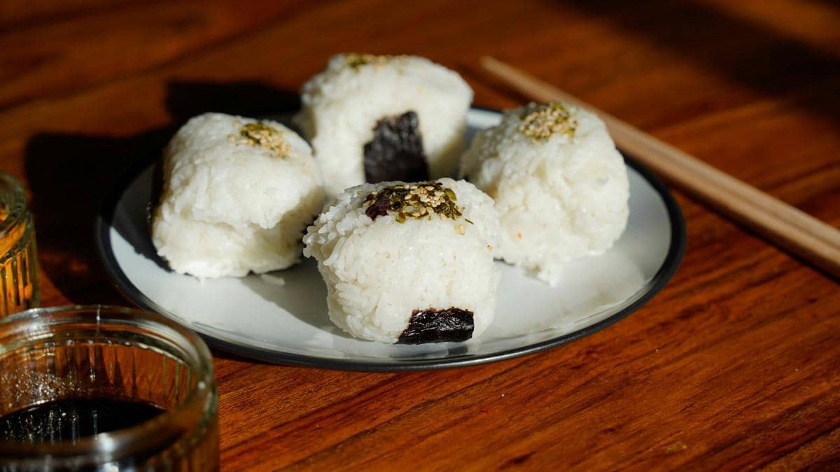 Beberapa onigiri nasi Jepang dengan taburan wijen dan rumput laut disajikan di atas piring putih di meja kayu dengan sumpit.
