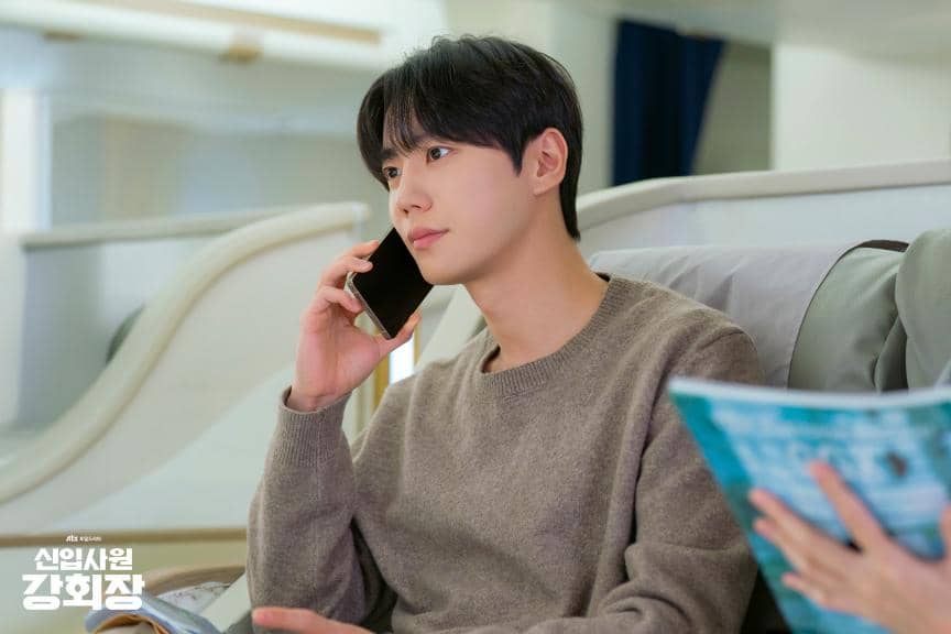 Seorang pria mengenakan sweater abu-abu sedang berbicara di telepon sambil duduk di kursi pesawat dan memegang majalah.