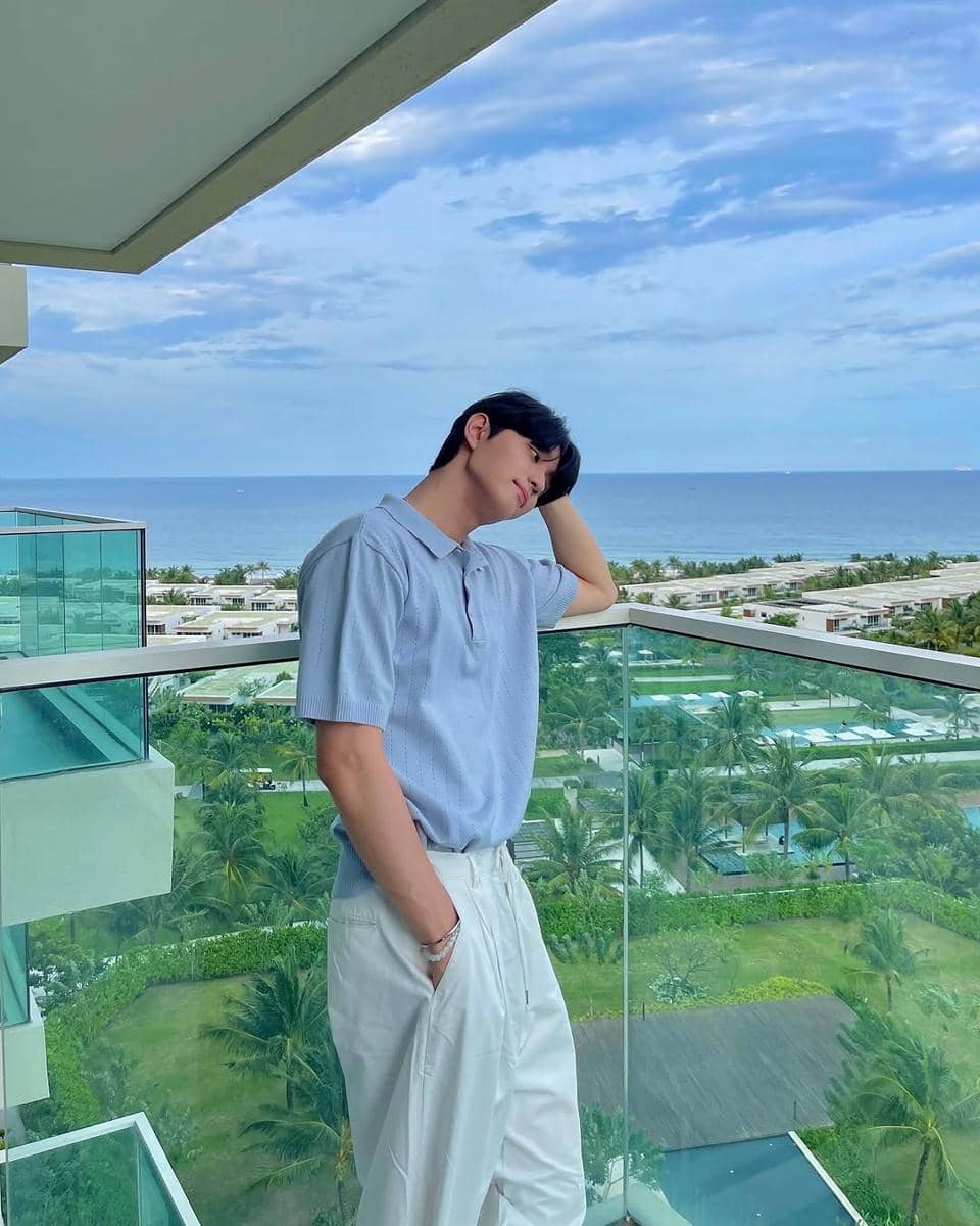 Pria berpose santai di balkon dengan pemandangan laut dan langit biru, mengenakan kemeja biru muda dan celana putih.