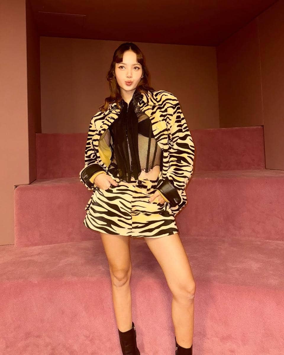 Seorang wanita berpose dengan gaya rambut medium length, mengenakan jaket dan rok bermotif zebra di ruangan bernuansa cokelat dan merah muda.