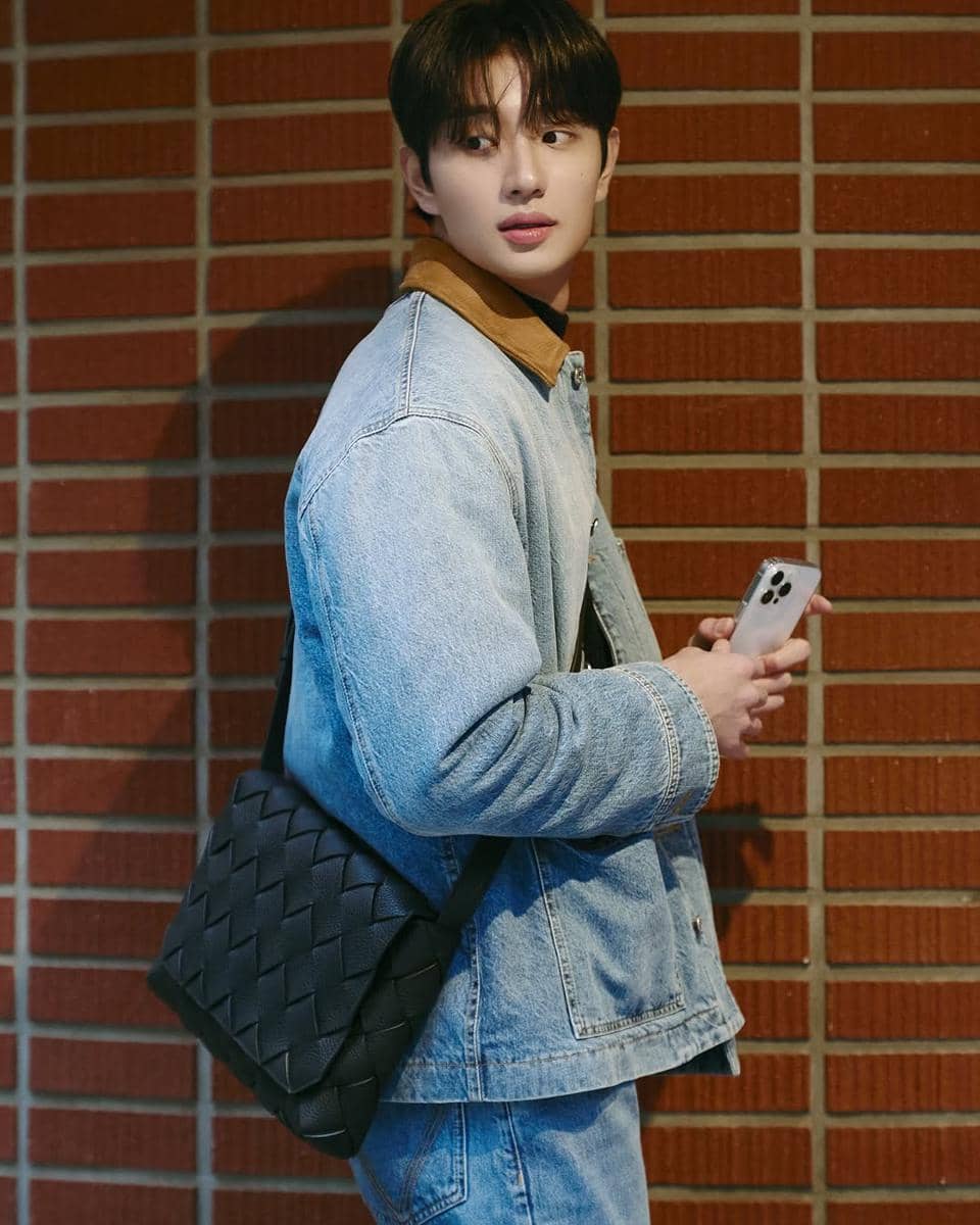 Seorang pria mengenakan jaket denim biru muda dan tas selempang hitam, memegang ponsel sambil menoleh di depan dinding bata merah.