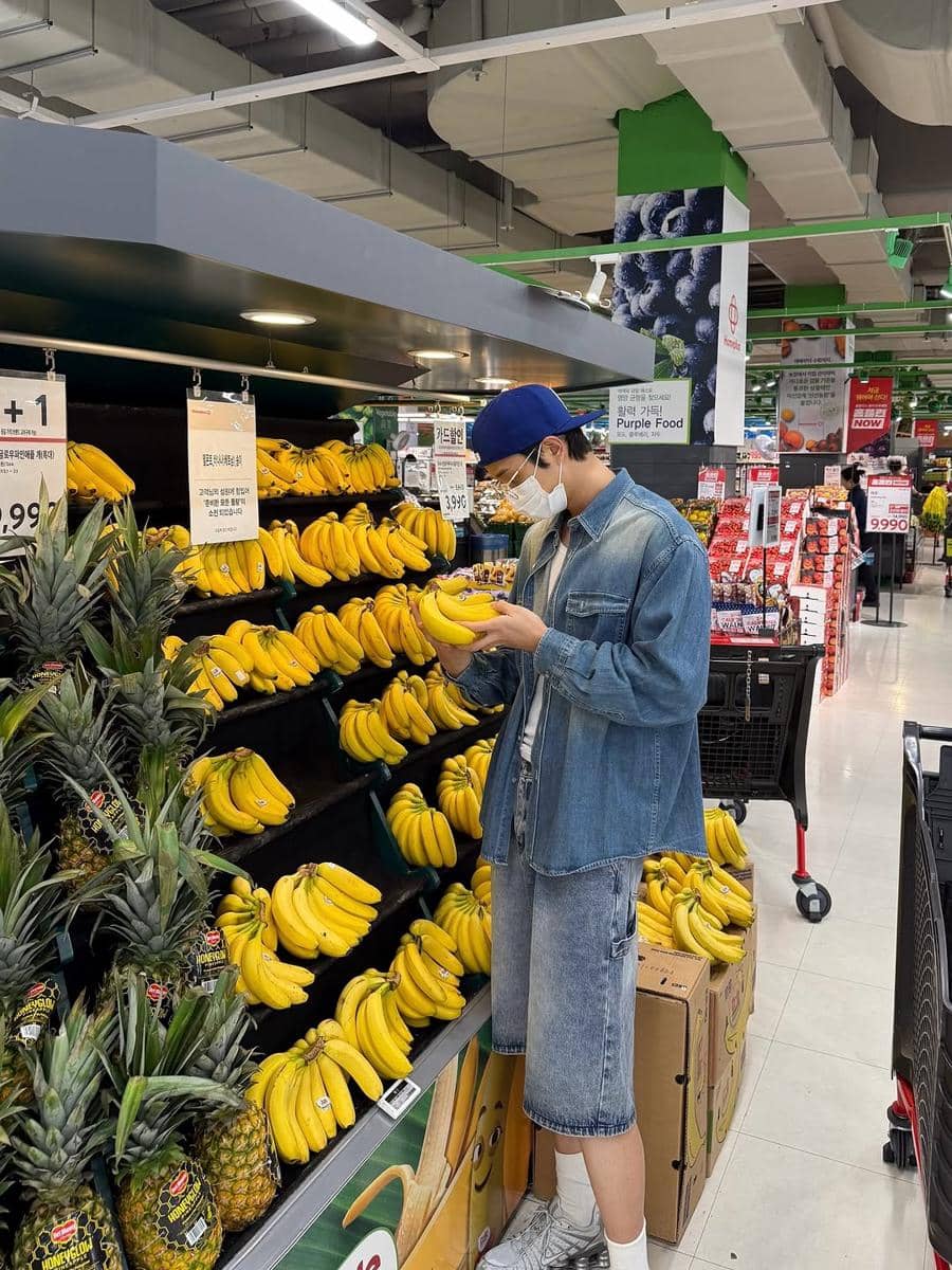 Pria mengenakan jaket denim, celana pendek jeans, dan topi biru sedang memilih pisang di bagian buah supermarket.