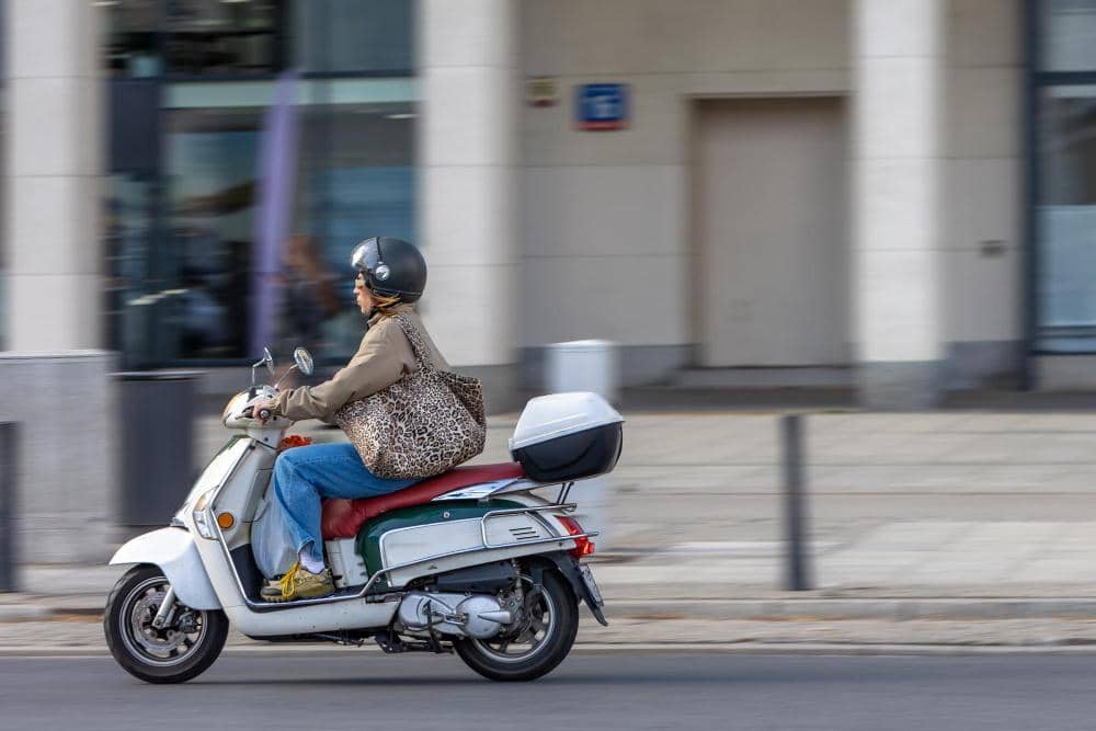 Seorang pengendara mengenakan helm dan jaket cokelat mengendarai motor listrik berwarna putih di jalan perkotaan.