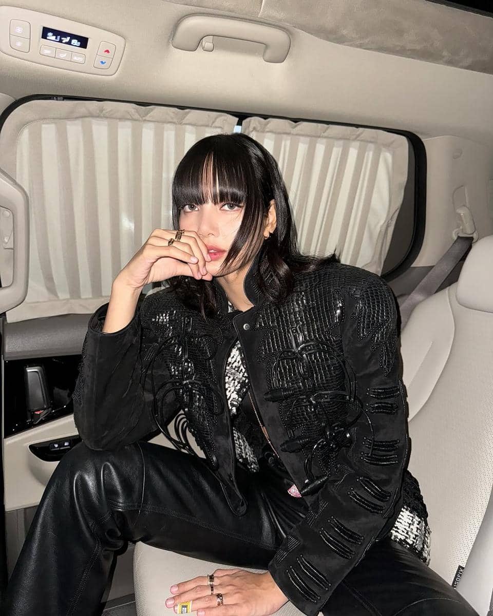 Seorang perempuan berambut medium dengan poni depan duduk di dalam mobil mengenakan jaket hitam dan celana kulit hitam.