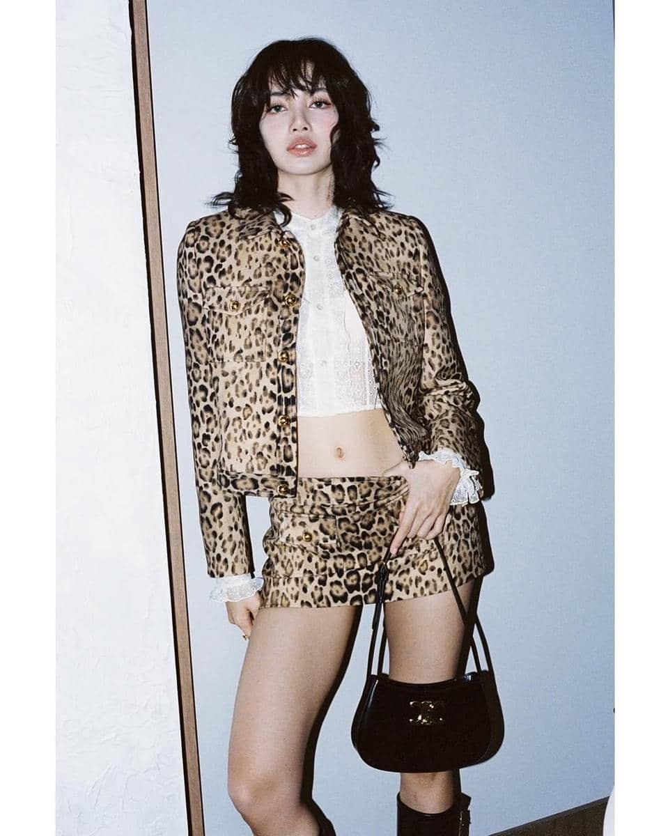 Seorang wanita berambut medium bergelombang mengenakan setelan motif leopard dengan rok mini dan jaket, memegang tas hitam.