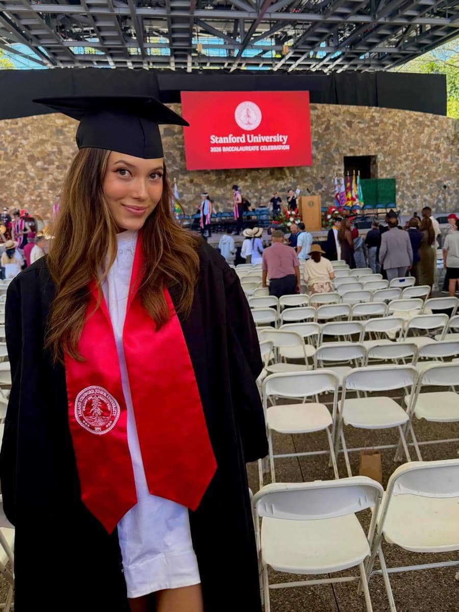 Seorang wisudawati mengenakan toga hitam dan selempang merah dengan latar belakang panggung bertuliskan Stanford University.