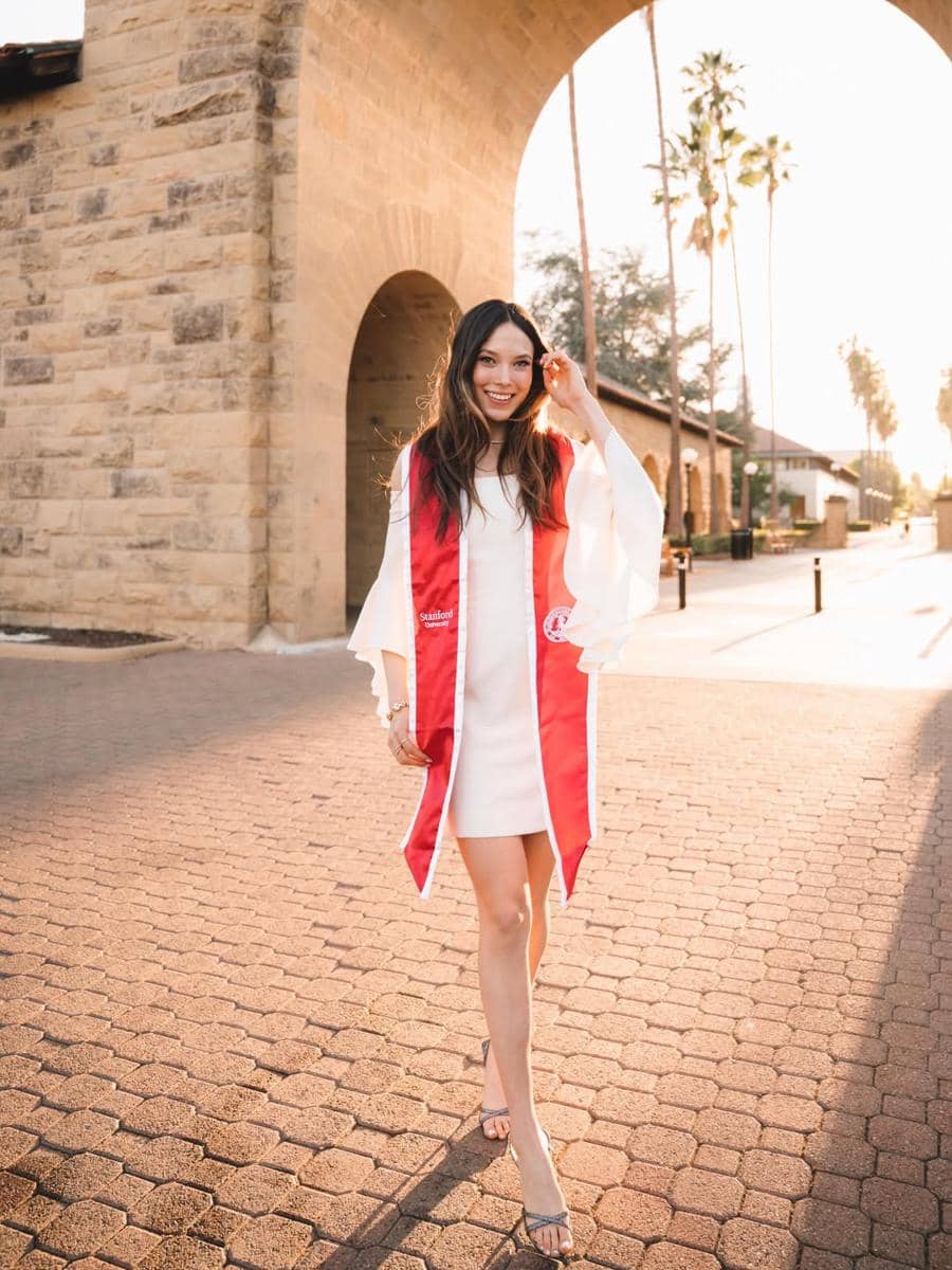 Seorang perempuan mengenakan gaun putih dan selempang merah berjalan di area kampus Stanford University saat matahari terbenam.