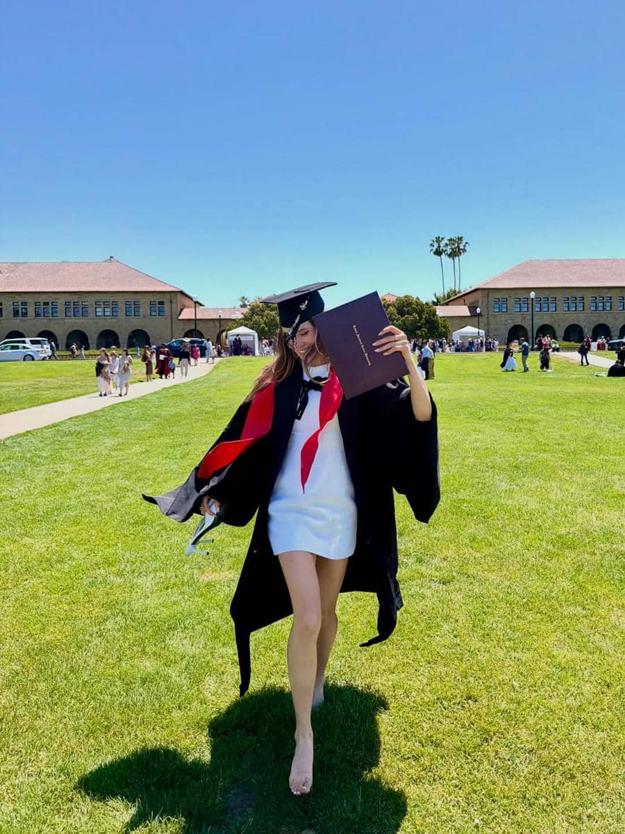 Seorang wisudawati mengenakan toga hitam dan gaun putih memegang ijazah di area kampus Stanford University pada hari cerah.