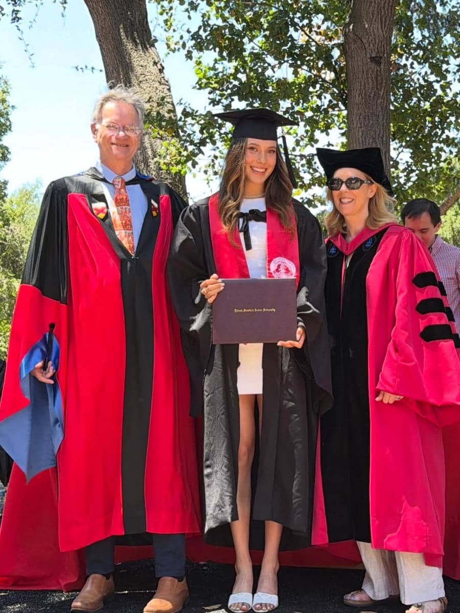 Seorang wisudawati mengenakan toga hitam dan syal merah berpose sambil memegang ijazah bersama dua dosen di acara kelulusan Stanford University.