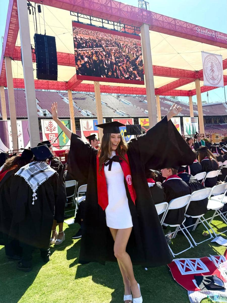 Seorang wisudawan mengenakan toga hitam dan syal merah berpose ceria di acara kelulusan di Stanford University.