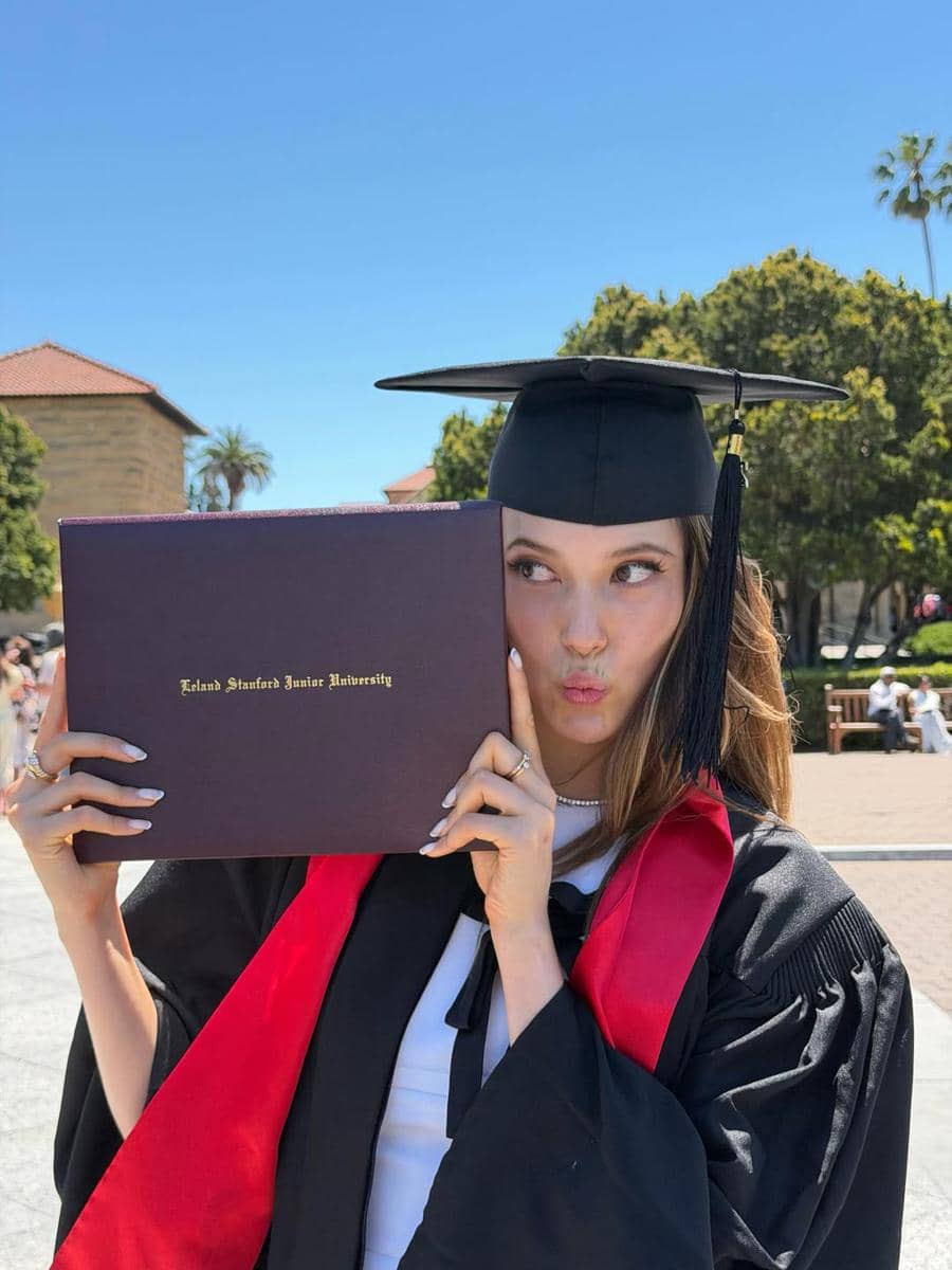 Seorang wisudawati mengenakan toga hitam dan selempang merah memegang ijazah Stanford University di luar ruangan pada hari cerah.