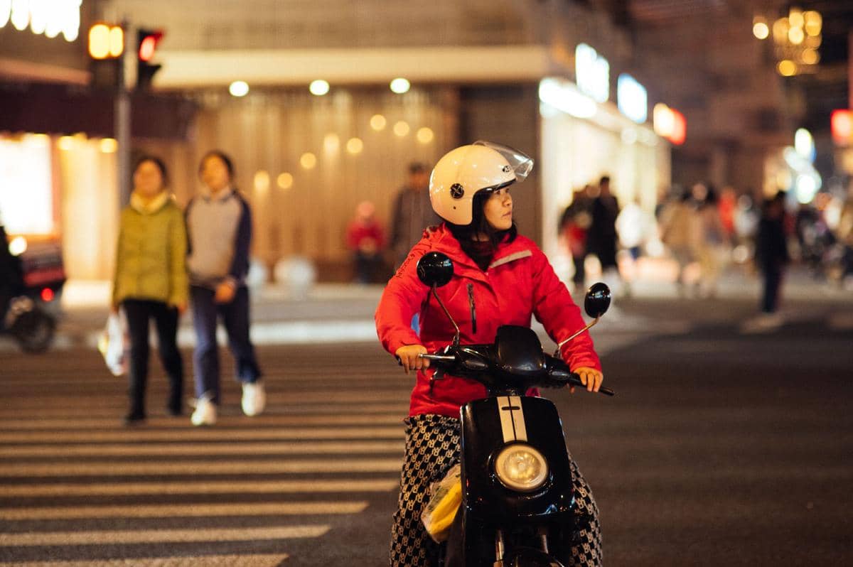 Seorang perempuan mengenakan jaket merah dan helm putih mengendarai motor listrik di jalan kota pada malam hari.