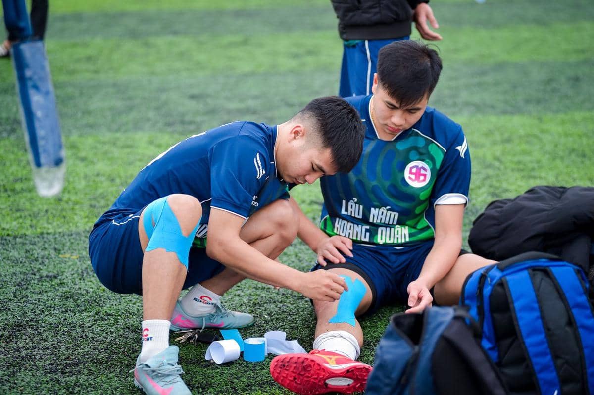 Dua pemain sepak bola mengenakan seragam biru sedang memasang perban atau tape biru di lutut di lapangan rumput sintetis.