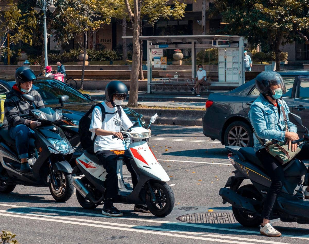 Beberapa pengendara mengenakan helm dan masker sedang berhenti di jalan dengan motor listrik di bawah sinar matahari.
