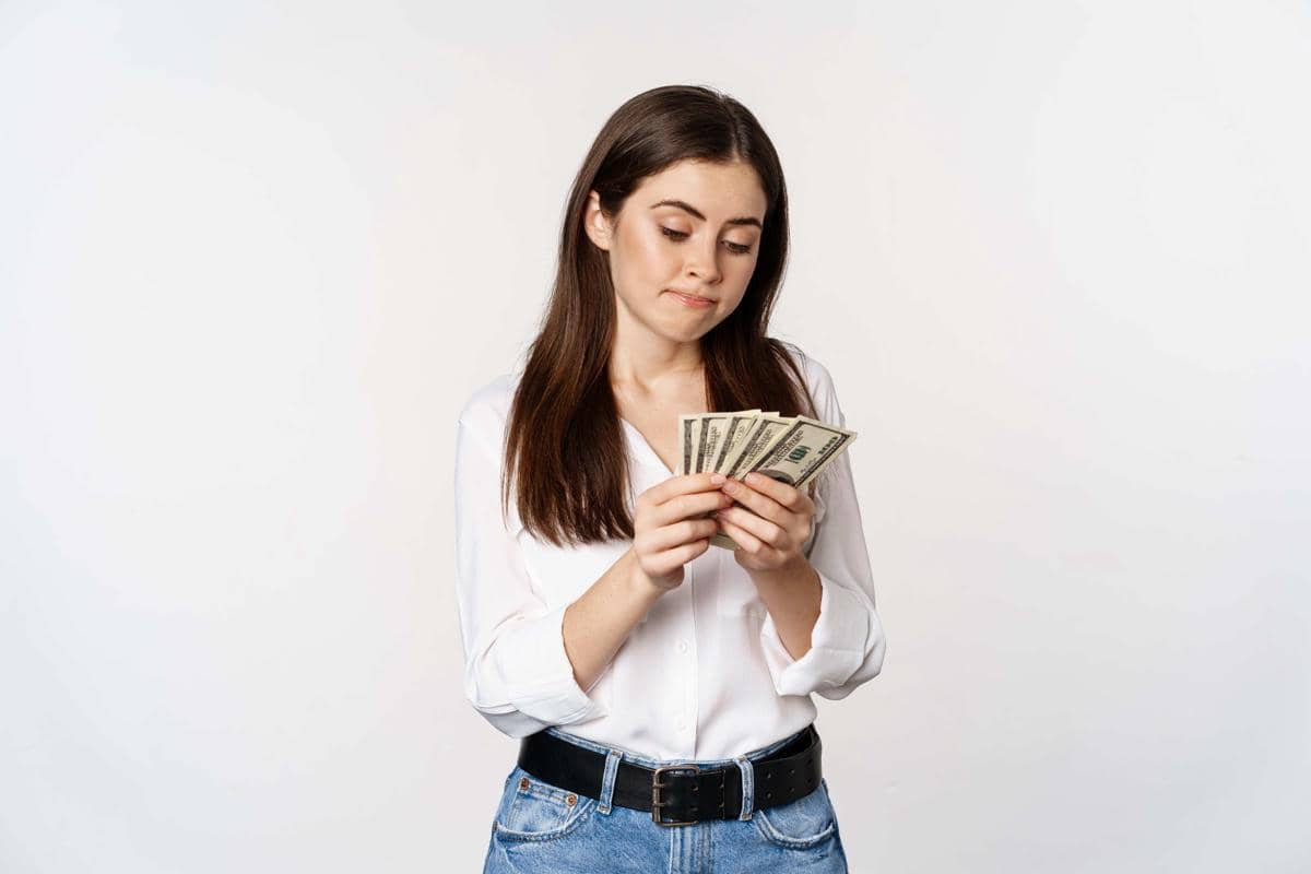 Seorang wanita muda mengenakan kemeja putih dan celana jeans sedang menghitung lembaran uang dolar di tangannya.