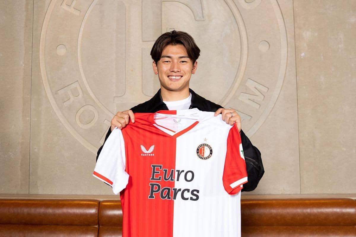 Ayase Ueda tersenyum sambil memegang jersey klub sepak bola Feyenoord berwarna merah dan putih di dalam ruangan.