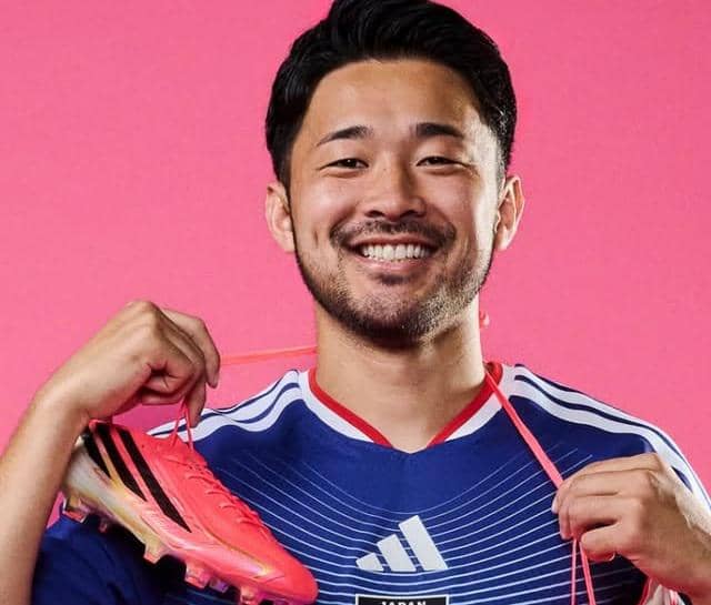 Yukinari Sugawara mengenakan jersey timnas Jepang berwarna biru sambil memegang sepatu bola berwarna merah muda