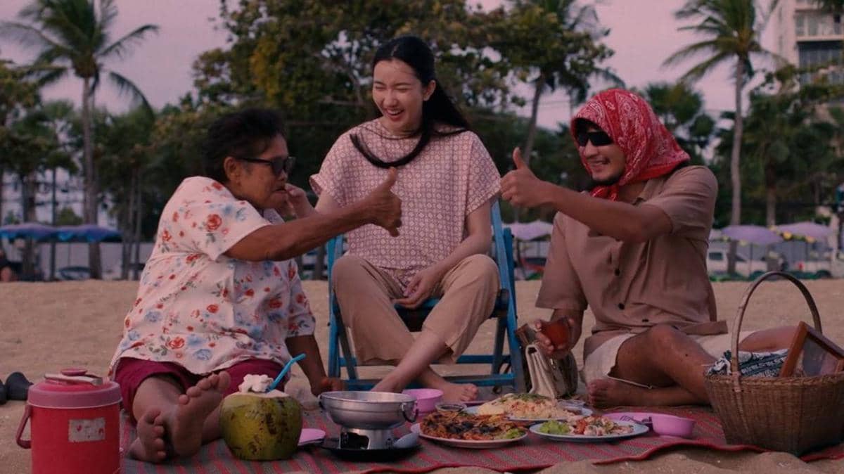 Tiga orang duduk santai di pantai sambil menikmati makanan dan minuman, tertawa bersama di suasana sore yang hangat.