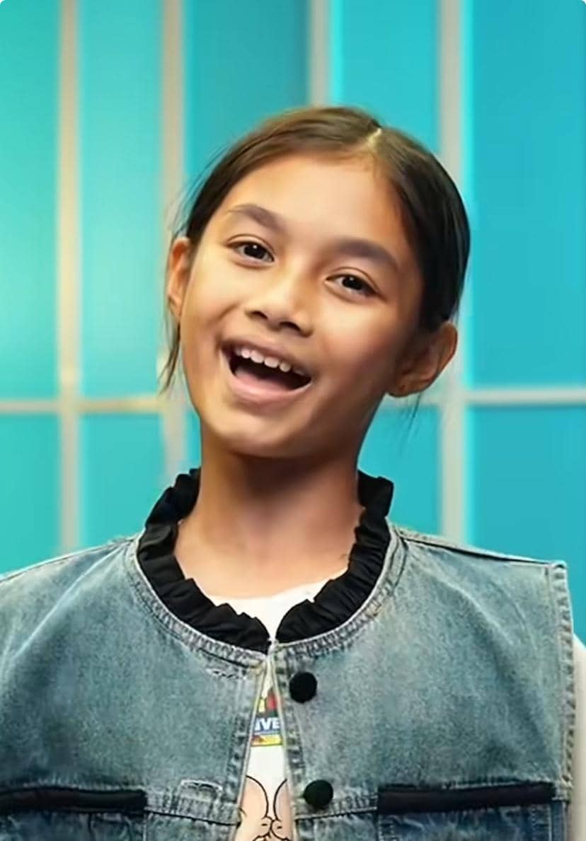 Seorang anak perempuan tersenyum mengenakan rompi denim biru dengan latar belakang berwarna biru muda bergaris vertikal.