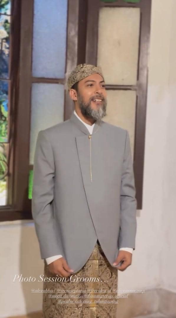 Seorang pria mengenakan beskap abu-abu dan blangkon batik berdiri tersenyum di depan jendela kaca berwarna saat sesi foto pernikahan.
