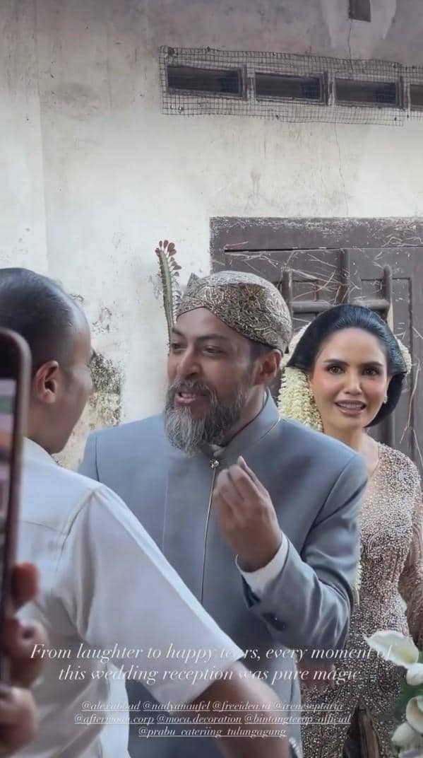 Pasangan pengantin mengenakan busana adat modern berwarna abu-abu dan keemasan sedang berbincang dengan tamu di resepsi pernikahan.