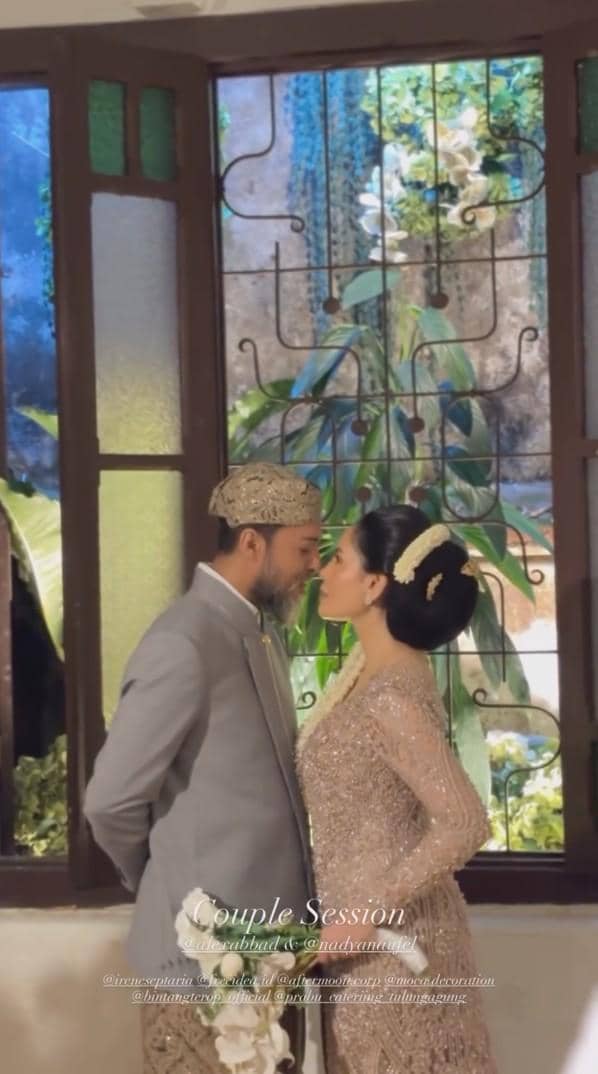 Pasangan pengantin mengenakan busana adat berwarna abu-abu dan keemasan saling menatap mesra di depan jendela kaca berwarna.