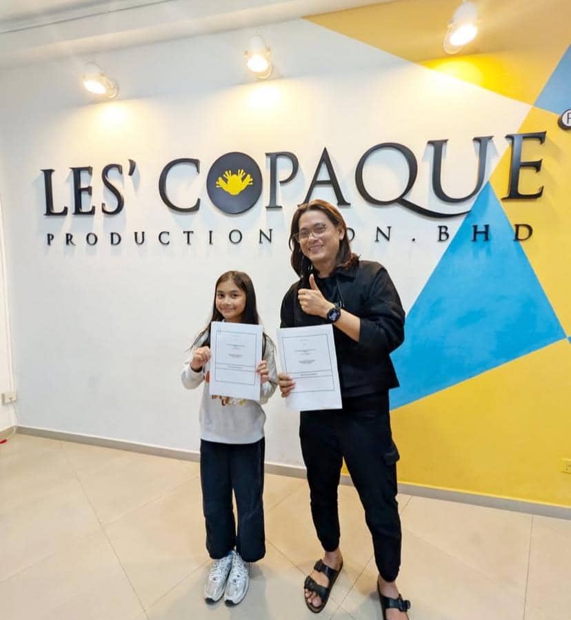 Seorang anak perempuan dan seorang pria berdiri di depan logo Les' Copaque Production sambil memegang dokumen dan tersenyum ke kamera.