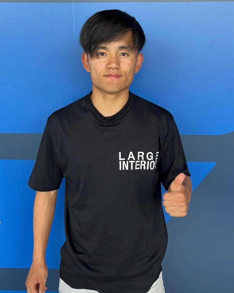 Takefusa Kubo mengenakan kaus hitam bertuliskan 'LARGE INTERIOR' berdiri di depan dinding biru sambil mengacungkan jempol.