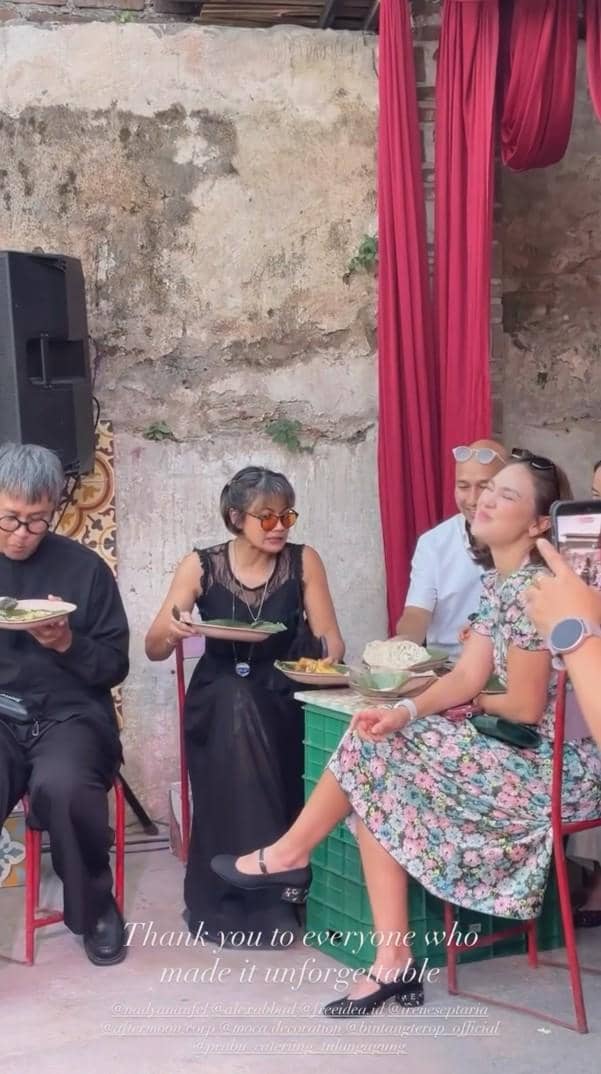 Tamu duduk menikmati hidangan di resepsi pernikahan dengan latar dinding bata dan dekorasi kain merah yang menjuntai.