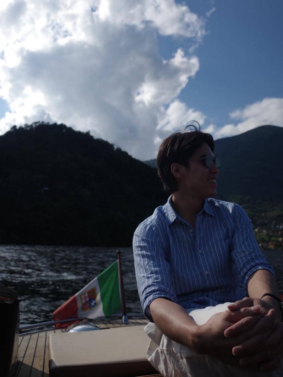 El Rumi duduk santai di atas perahu di Danau Como dengan latar pegunungan dan bendera Italia di belakangnya.
