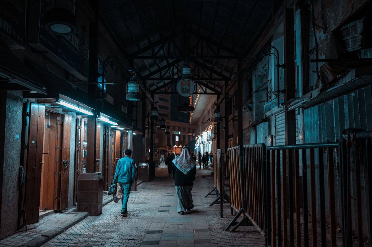 Lorong kota pada malam hari dengan pencahayaan biru dan oranye, menampilkan beberapa orang berjalan di antara bangunan tua.