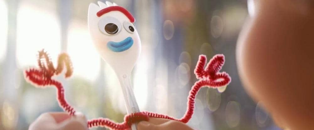 Karakter mainan Forky dari film animasi Toy Story 4 dipegang oleh tangan anak kecil dengan latar belakang buram berwarna cerah.