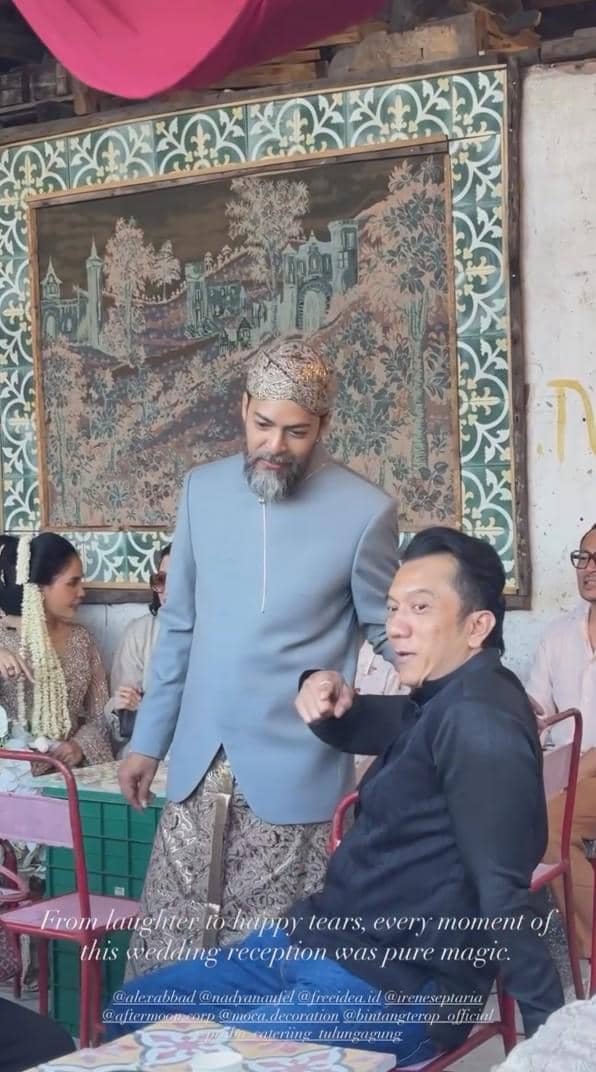 Suasana resepsi pernikahan dengan tamu berpakaian adat, salah satu pria mengenakan beskap abu-abu dan blangkon berbincang dengan tamu lain.
