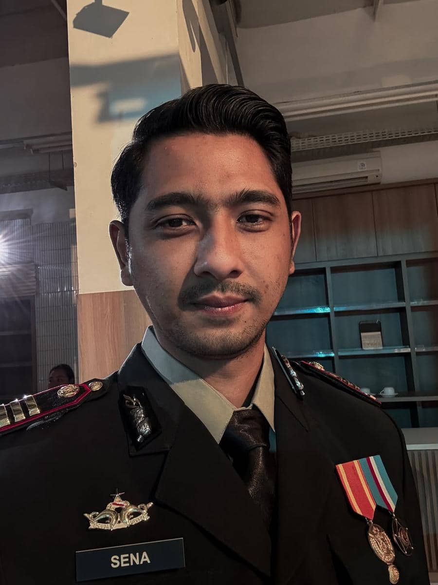 Seorang pria mengenakan seragam dinas lengkap dengan tanda pangkat dan medali, berdiri di dalam ruangan dengan pencahayaan hangat.