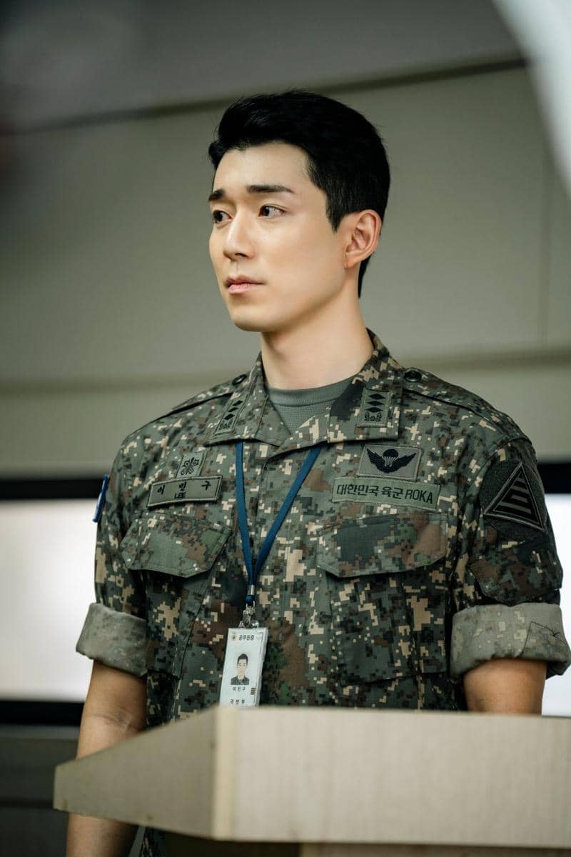 Seorang pria mengenakan seragam militer Korea berdiri di belakang podium dengan ekspresi serius dalam adegan drama The Legend Of Kitchen Soldier.