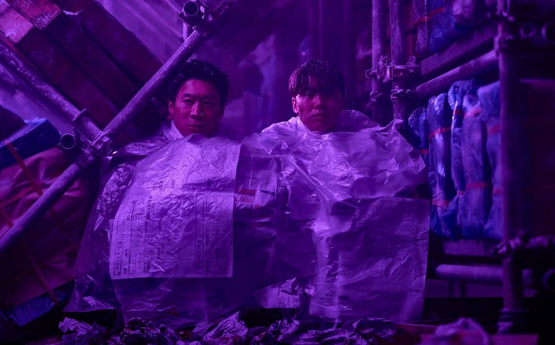 Dua pria bersembunyi di antara tumpukan barang dan pipa logam dengan pencahayaan ungu dalam cuplikan film Husbands in Action.