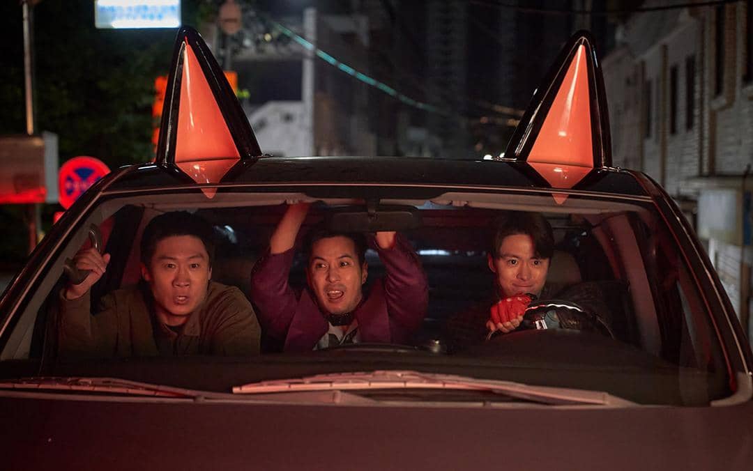 Tiga pria duduk di dalam mobil pada malam hari dengan ekspresi tegang dan bersemangat, dua kerucut oranye besar terlihat di atap mobil.