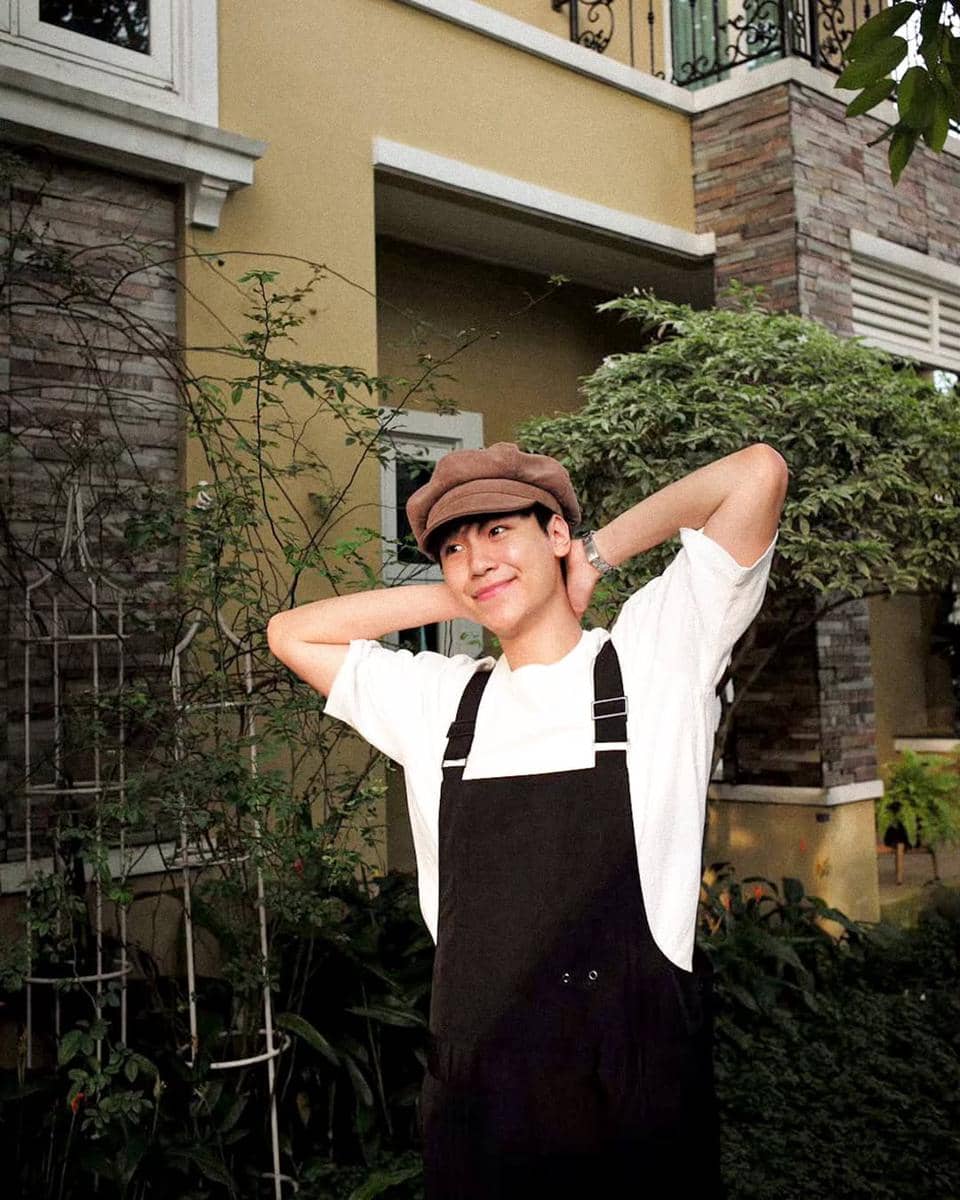 Seorang pria mengenakan kaus putih dan overall hitam dengan topi cokelat berpose santai di halaman rumah bergaya modern.