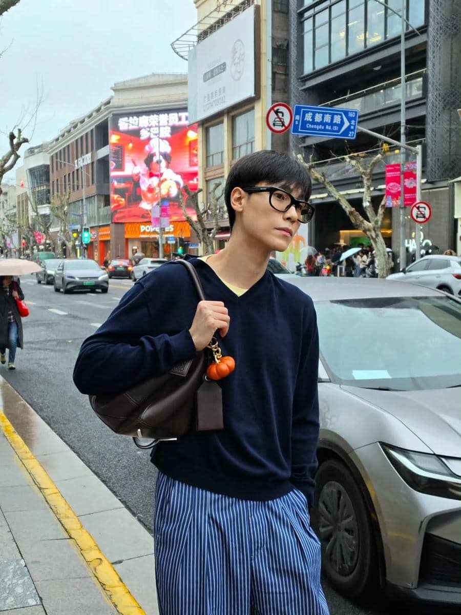 Seorang pria bergaya kasual mengenakan sweater biru tua dan celana garis-garis biru di jalan kota dengan suasana modern dan ramai.
