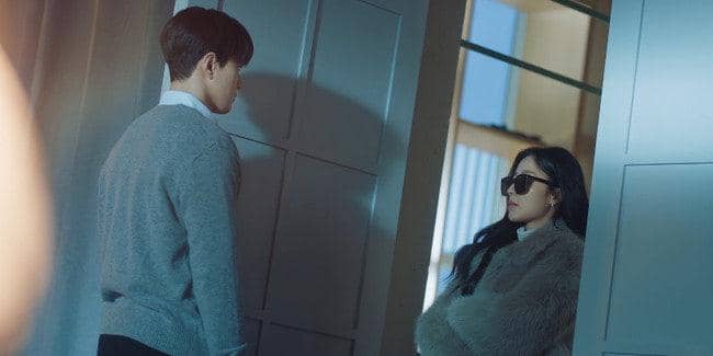 Seorang pria mengenakan sweater abu-abu berdiri di depan seorang wanita berjaket bulu dan berkacamata hitam di dalam ruangan modern.