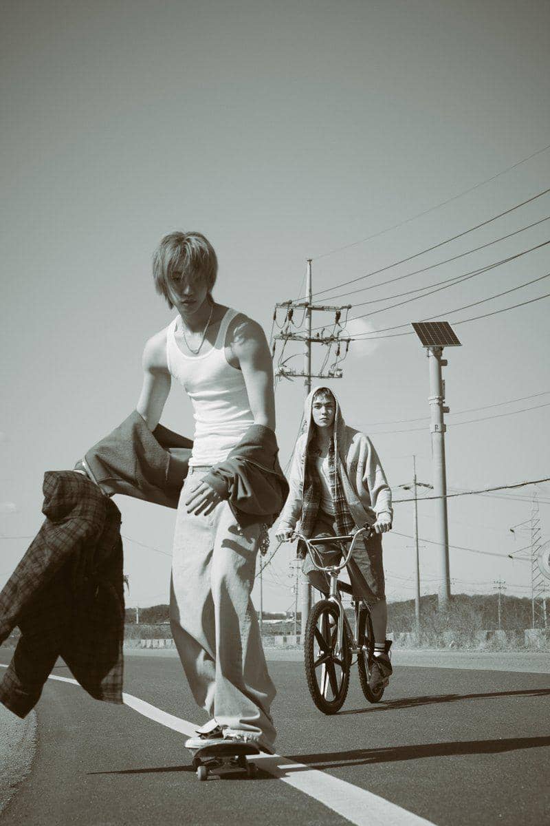 Dua pria muda bergaya kasual sedang beraktivitas di jalan, satu mengendarai skateboard dan satu lagi bersepeda di bawah langit cerah.