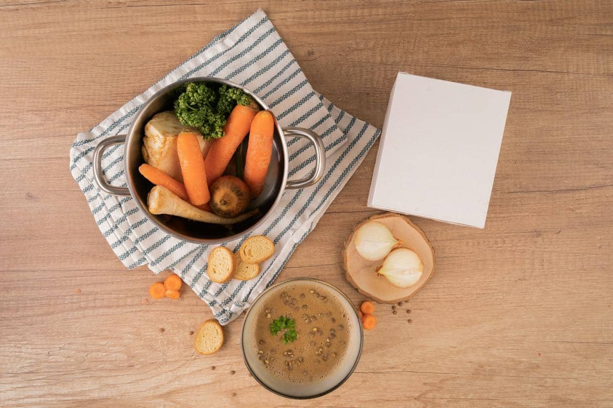 Bahan-bahan untuk membuat sup seperti wortel, brokoli, bawang, dan roti kering tersusun di atas meja kayu dengan panci dan mangkuk sup.
