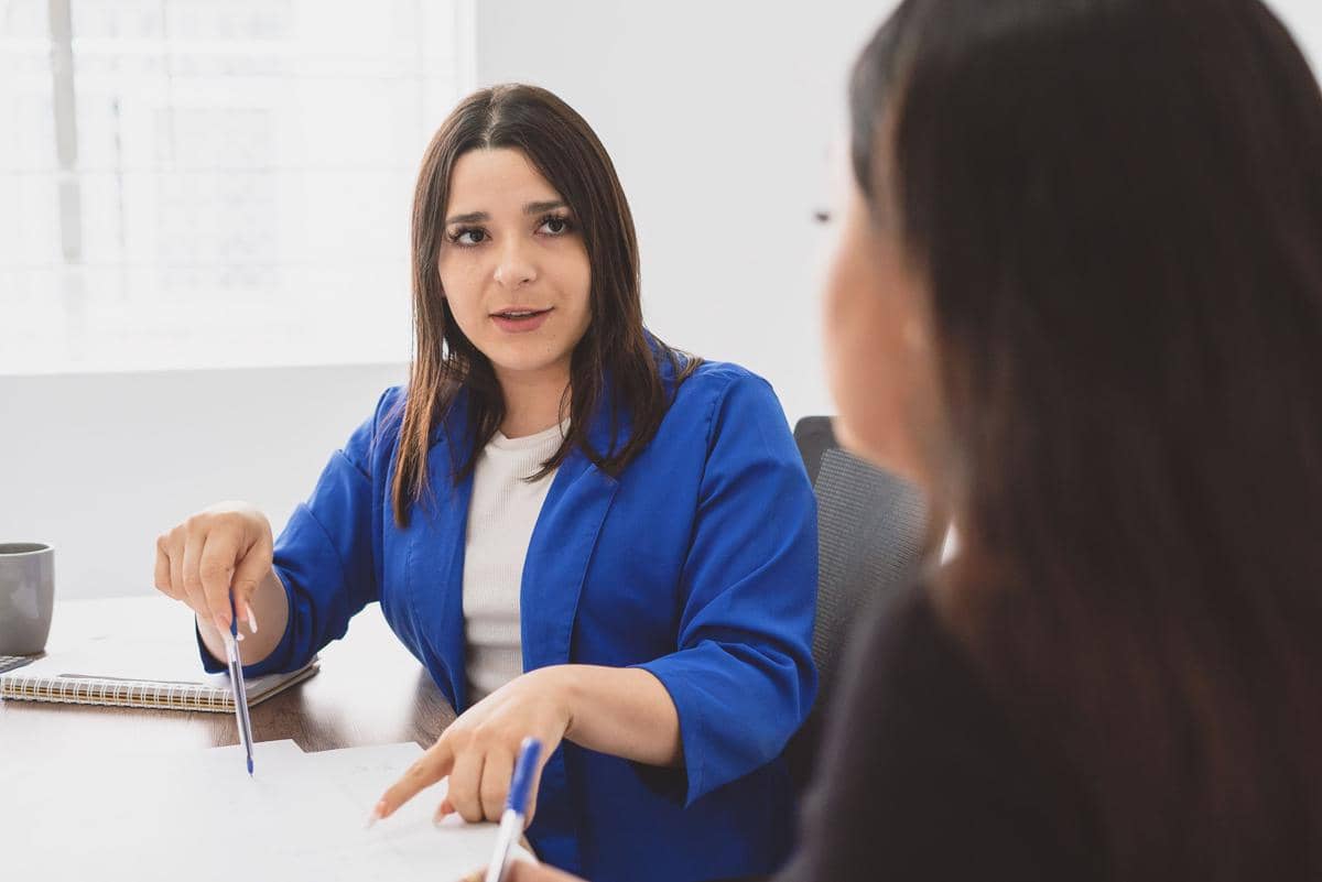 Dua wanita sedang berdiskusi di meja kantor, salah satunya mengenakan blazer biru sambil menunjuk dokumen dengan pena.