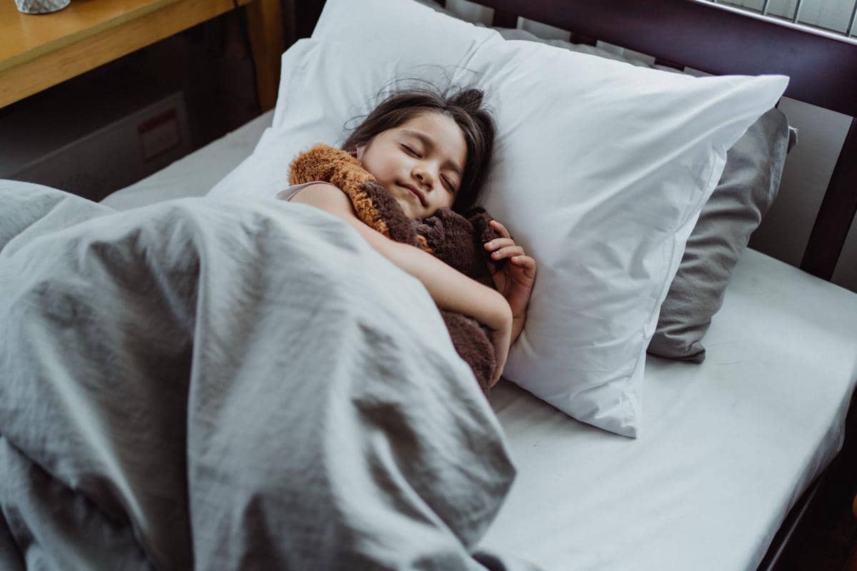 Seorang anak tidur nyenyak di atas tempat tidur, berbaring di bawah selimut dalam suasana kamar yang tenang.