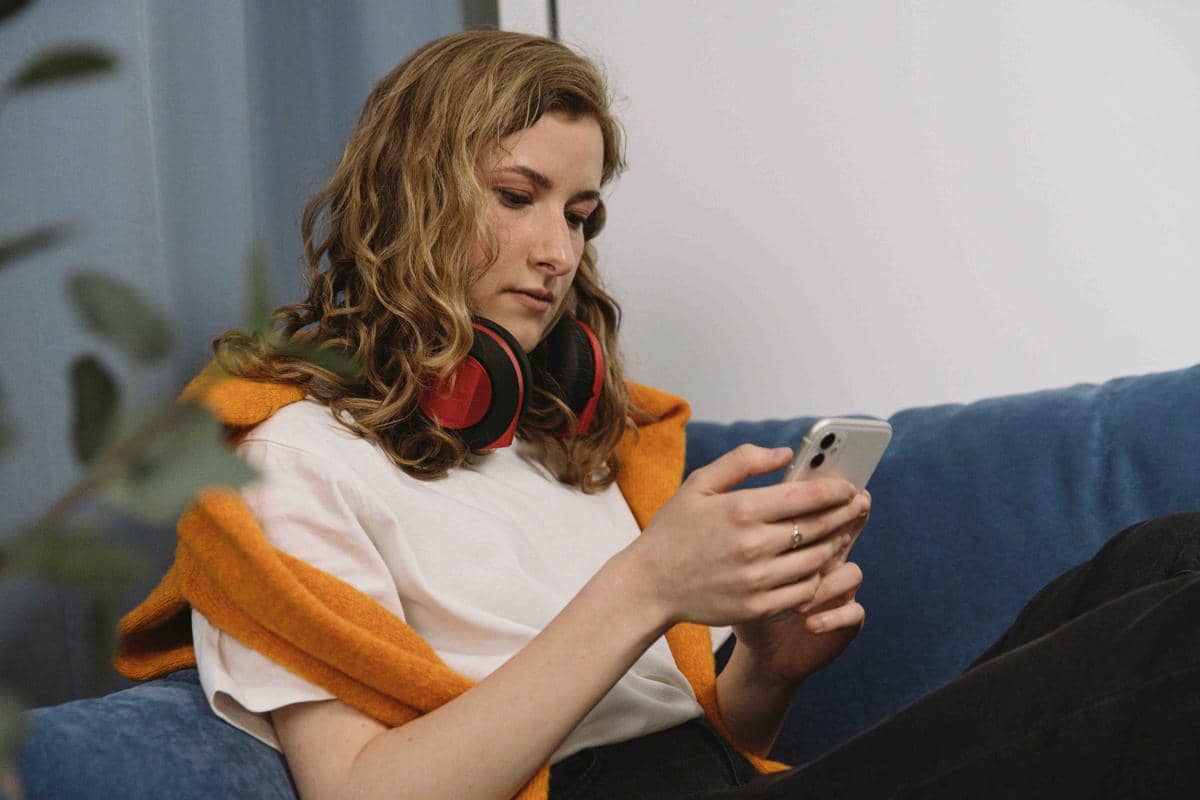 Seorang perempuan duduk di sofa biru sambil memegang ponsel, mengenakan headphone besar di leher dan sweter oranye di bahu.
