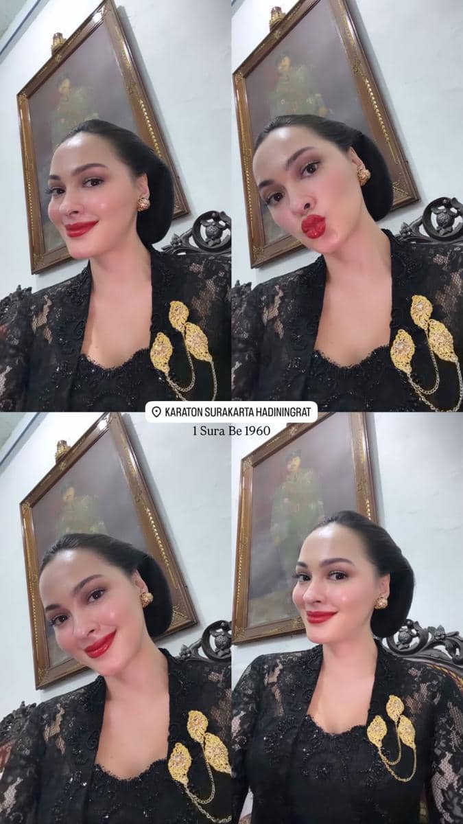 Seorang perempuan mengenakan kebaya hitam berhias bros emas berpose di dalam ruangan dengan lukisan tokoh berpakaian adat di dinding.