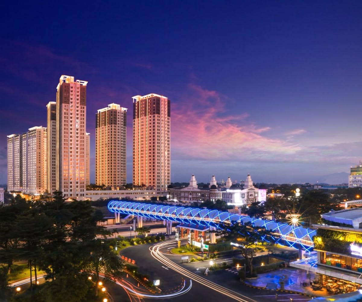 Pemandangan malam kawasan Gading Serpong dengan gedung-gedung tinggi dan jembatan penyeberangan bercahaya biru di bawah langit senja.