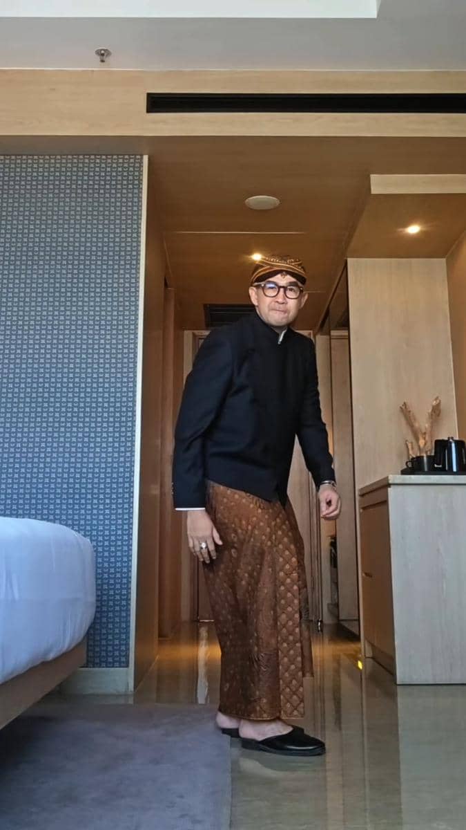 Seorang pria mengenakan beskap hitam, jarik batik cokelat, dan blangkon berdiri di dalam kamar hotel dengan pencahayaan hangat.